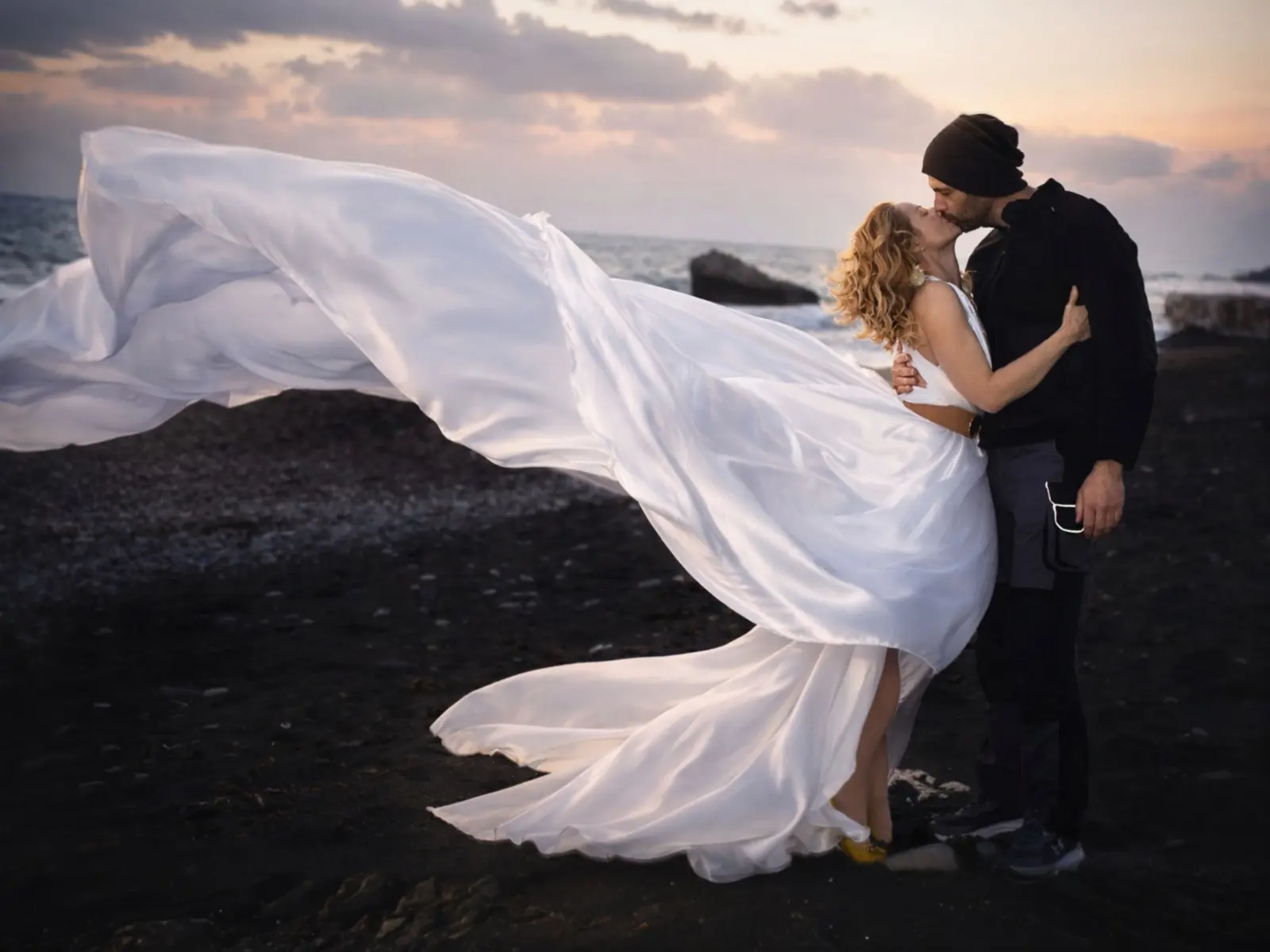 Bride and groom kissing on a Paphos beach at dusk while her silk gown flies in the wind
